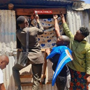 Photograph showing four people installing a red house number plate labeled "MY/A/006/A" above a doorway covered with a patterned cloth. The scene highlights community effort in marking or identifying a residence, with individuals actively reaching up to secure the plate against a corrugated metal structure.